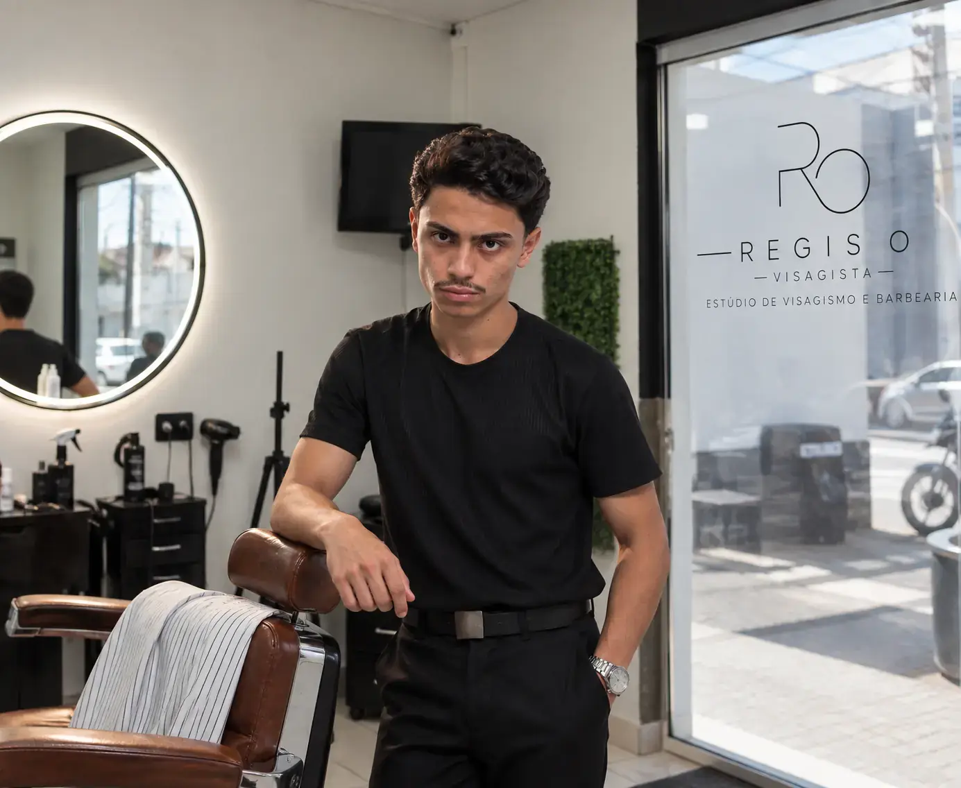 Regis O., visagista, de camiseta preta, apoiado na cadeira do seu estúdio em Passos